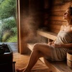 Kobieta owinięta ręcznikiem siedzi w drewnianej saunie obok pieca z rozgrzanymi kamieniami