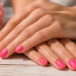 Dłonie z różowym manicure hybrydowym ułożone na drewnianym blacie obok różowych kwiatów